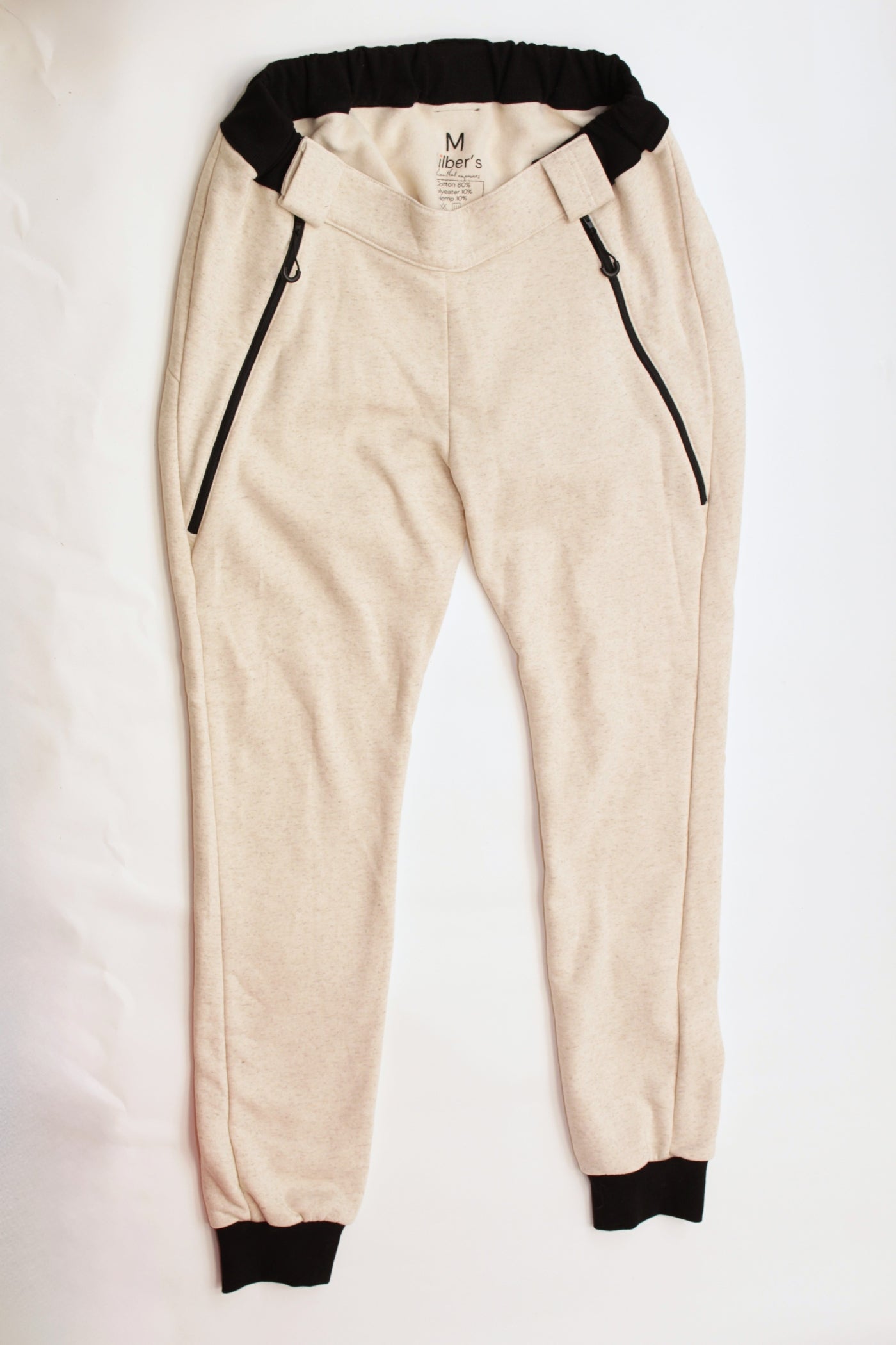 Here is the alt text you requested:
Eine beigefarbene Jogginghose mit schwarzen Bündchen an Bund und Knöcheln sowie schwarzen Reißverschlusstaschen, flach ausgelegt auf einem weißen Hintergrund. Es ist eine adaptive Rollstuhlhose von Vilber's, die speziell für Menschen mit Behinderungen entwickelt wurde.