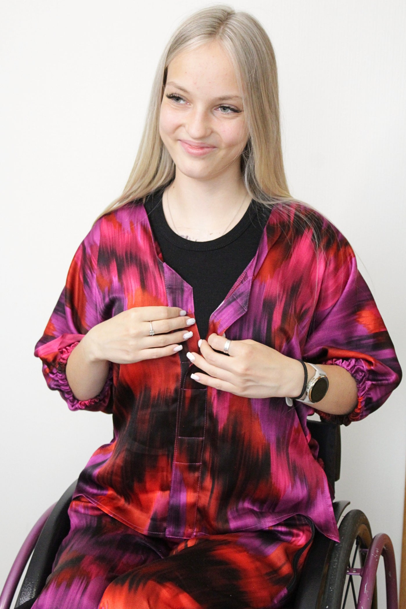 Ein langes, blondes Model mit einem leichten Lächeln trägt einen luxuriösen zweiteiligen Satz aus 100 % Viskose-Satin in leuchtenden Farben wie Fuchsia, Lila und Orange. Das Set ist für Rollstuhlfahrer geeignet und verfügt über magnetische Verschlüsse, was es auch für Menschen mit eingeschränkter Handfunktion, wie Tetraplegiker, leicht an- und auszuziehen macht.