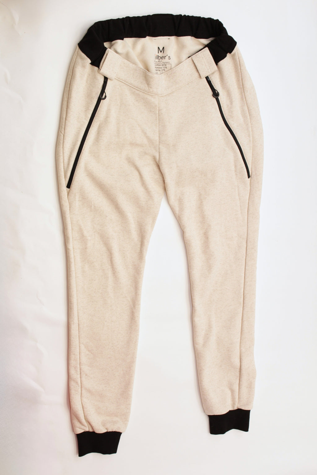 Here is the alt text you requested:

Eine beigefarbene Jogginghose mit schwarzen Bündchen an Bund und Knöcheln sowie schwarzen Reißverschlusstaschen, flach ausgelegt auf einem weißen Hintergrund. Es ist eine adaptive Rollstuhlhose von Vilber's, die speziell für Menschen mit Behinderungen entwickelt wurde.