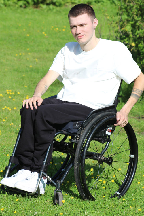 Young man using a manual wheelchair outdoors, wearing Vilber's adaptive joggers in black. Side view showing the seated fit and clean silhouette.