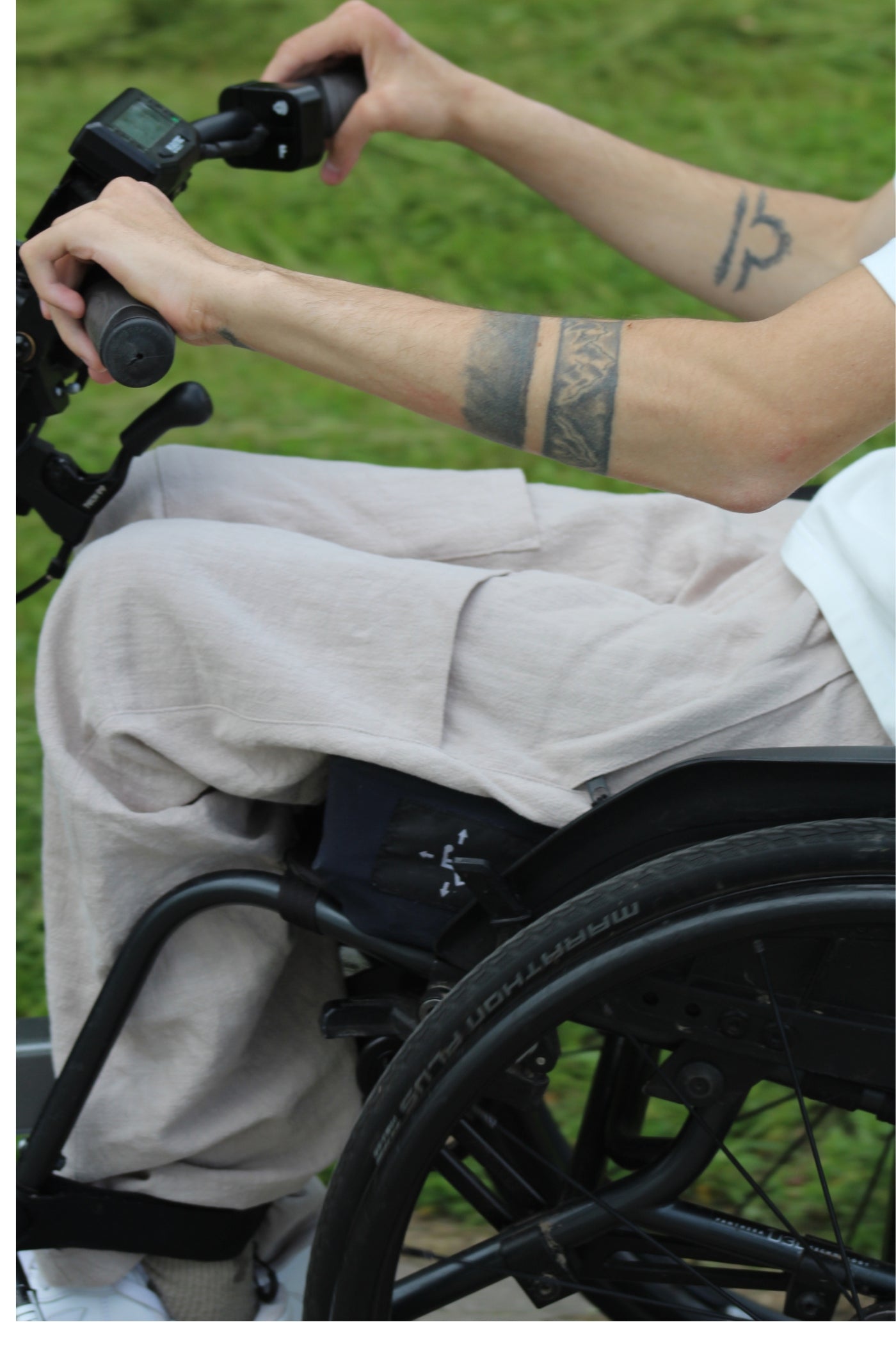 Close-up of a wheelchair user's hands on a Batec power add-on handlebar, with Vilber's beige summer trousers visible across the lap.